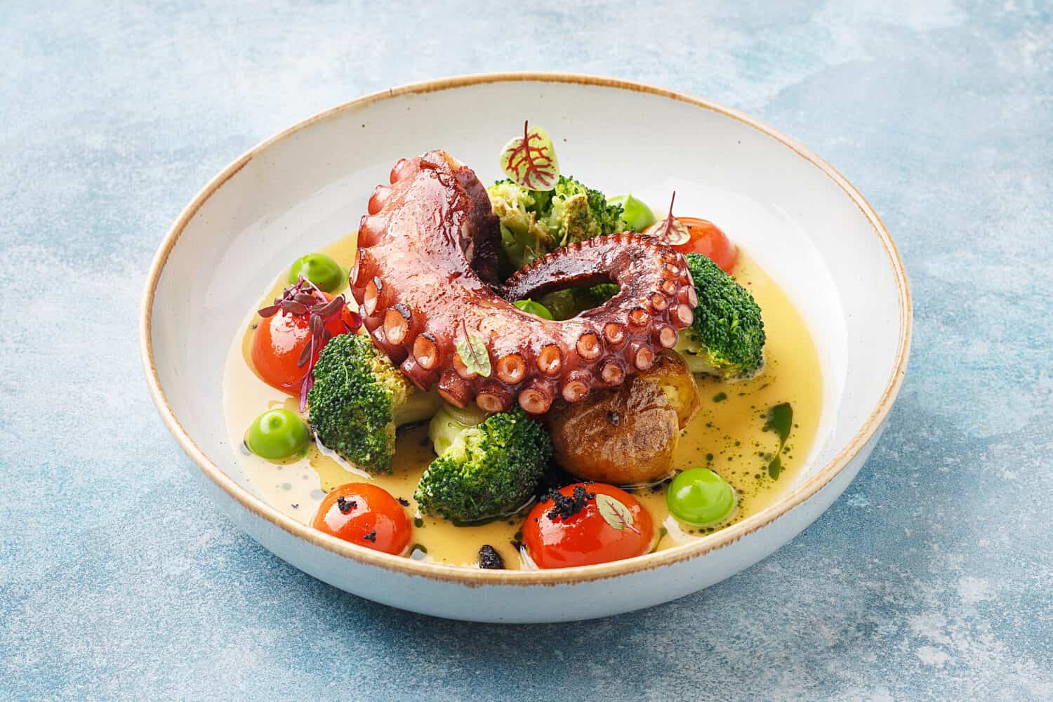 Grilled octopus tentacle with broccoli, cherry tomatoes and baked baby potatoes in white porcelain plate over blue concrete background. Side view, close up