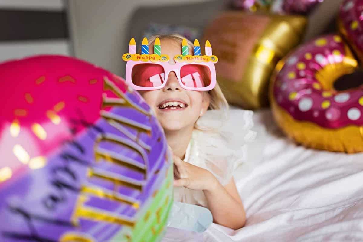 Indoor shot of pretty joyful cute adorable girl kid celebrating eight years old birthday with bright and colorful balloons with inscription words stay fabulous, wearing casual fashionable dress