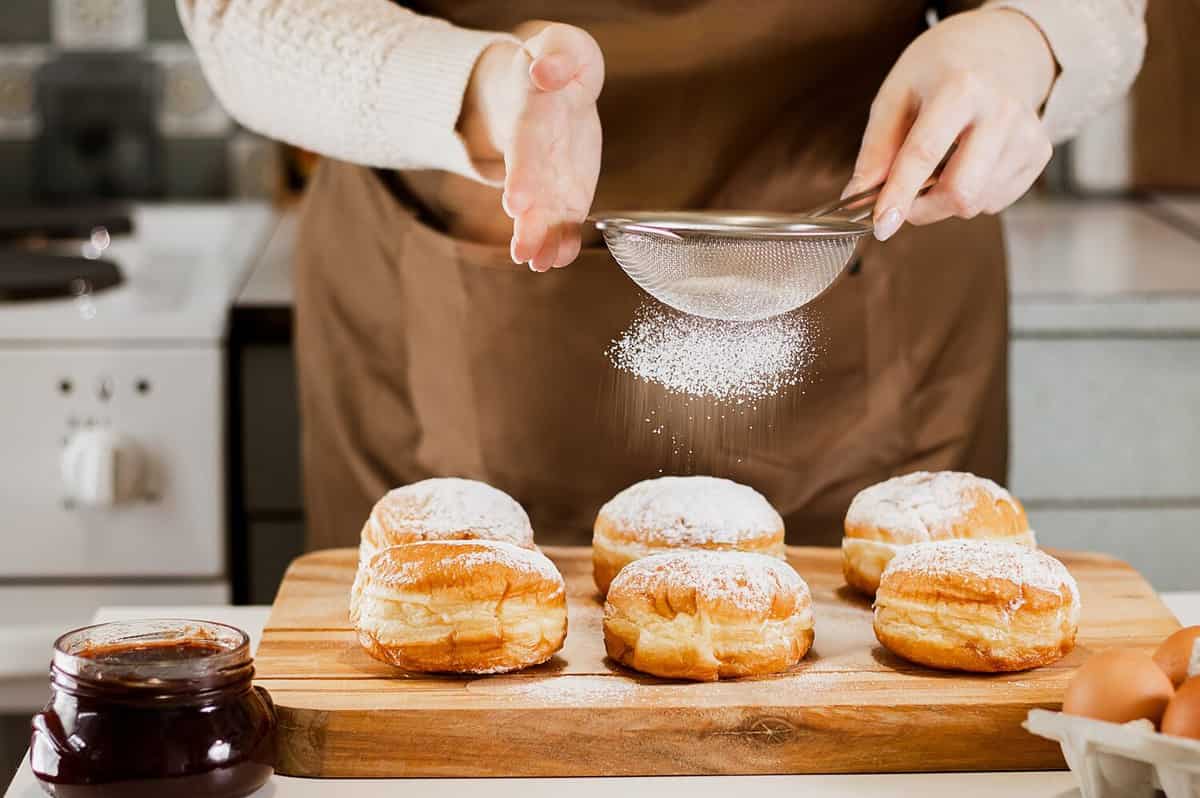 Woman prepares fresh donuts with jam in home kitchen. Cooking traditional Jewish Hanukkah sufganiyot. Hands sprinkle Berliners with powdered sugar.