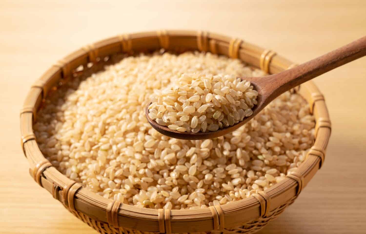 Brown rice in a bamboo colander and a wooden spoon on a wooden background. A pile of brown rice.