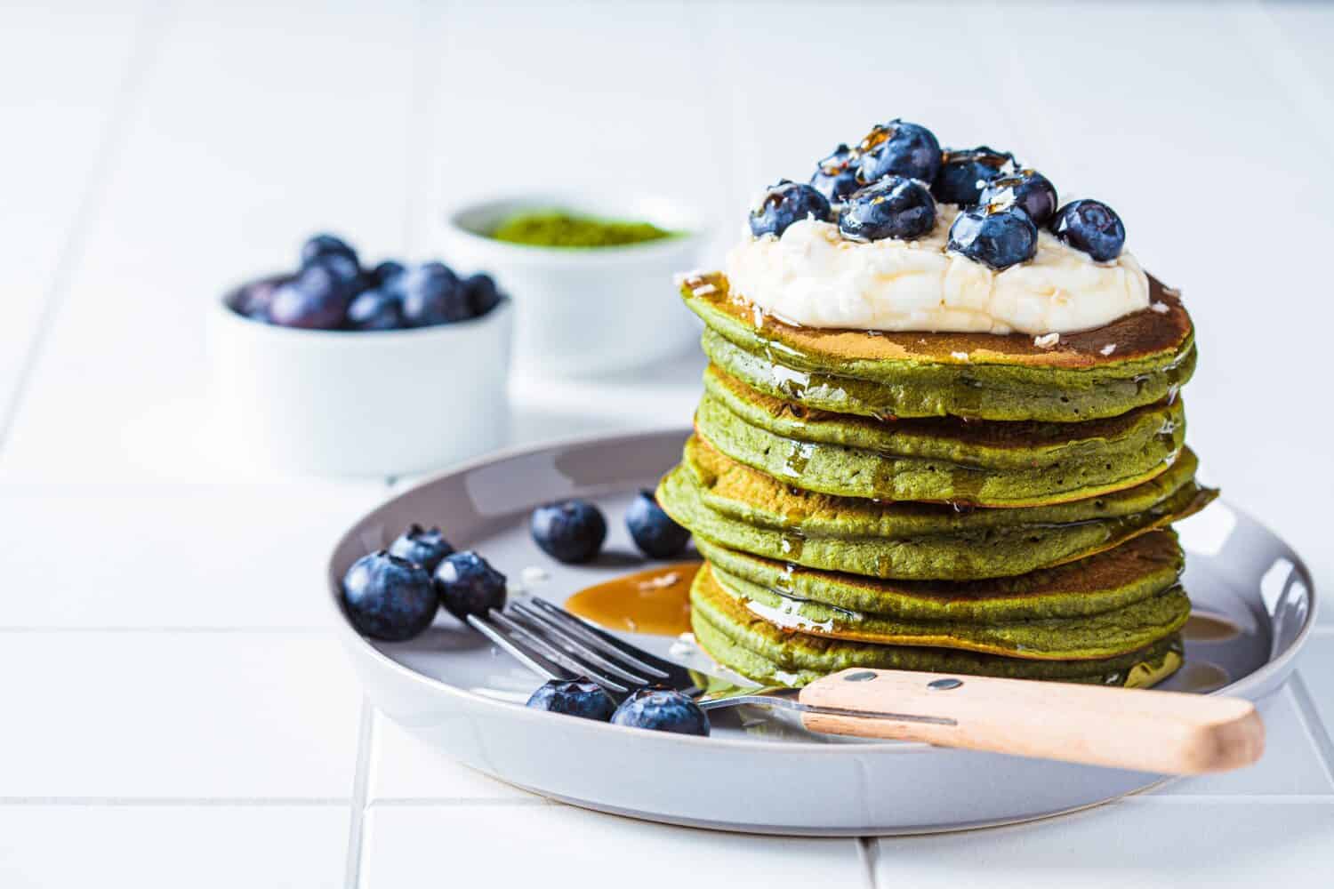 Matcha tea green pancakes with coconut cream, blueberries and maple syrup, white background, copy space. Vegan dessert concept.