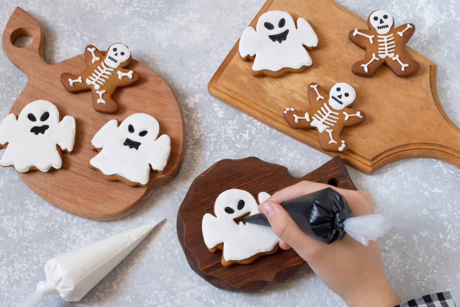 Decorating Halloween gingerbreads of ghosts and skeletons with frosting. Girl holds pastry bag with black icing and decorates gingerbread ghost