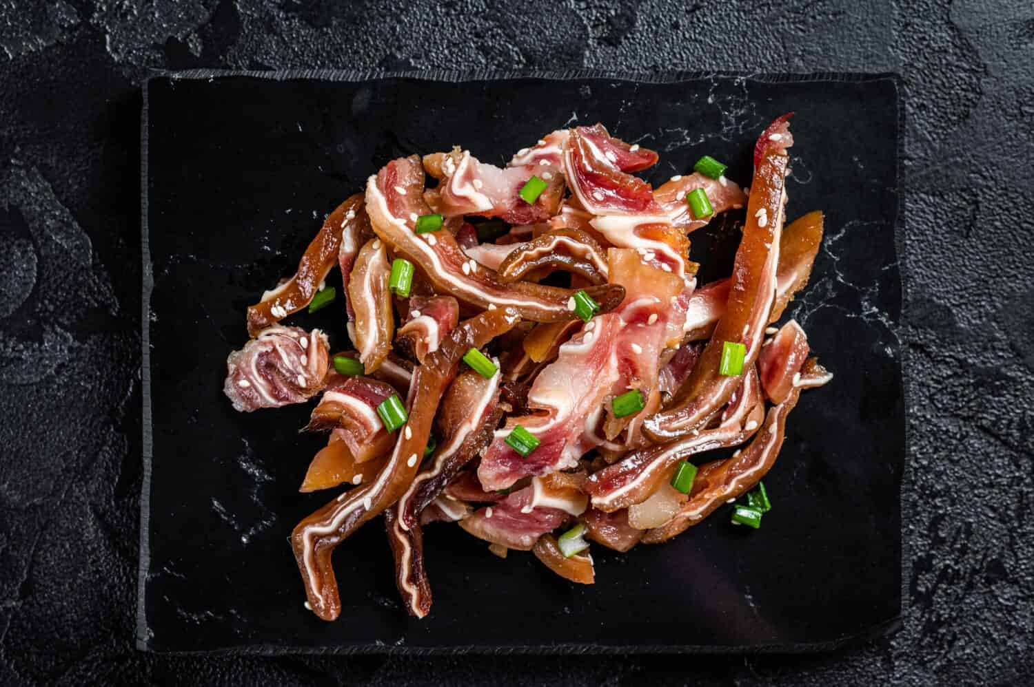 Fried pig ears with sesame seeds and green onions. Fried pork ears in korean style with spicy sauce. Black background. Top view.