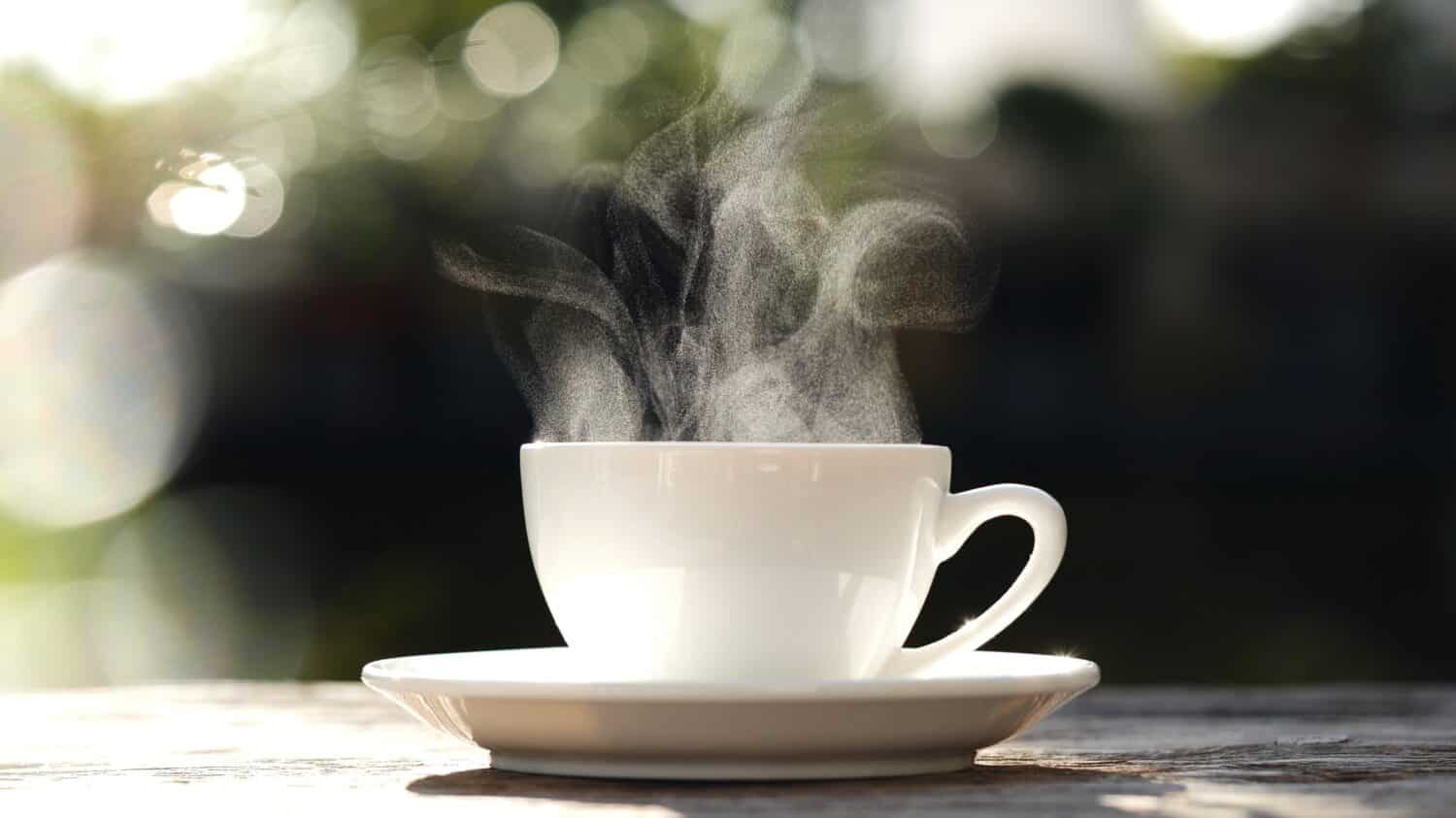 White coffee cup, mug with steaming smoke of coffee on old wooden table in morning nature outdoor, garden background, 4K. Hot Coffee Drink, Beverage Concept.