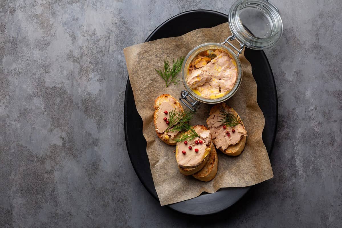 Plate with baguette toast with foie gras pate, directly above. A specialty food product made of the liver of a duck or goose, in a glass jar. Decorated with red pepper and dill.