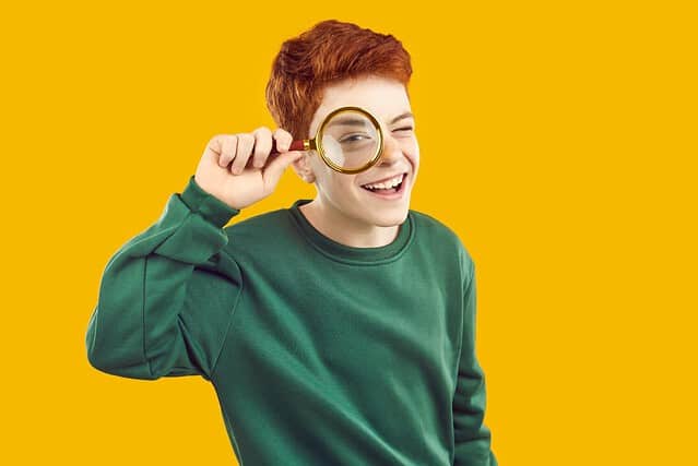 Children's interest. Portrait of cheerful active preteen boy who looks at camera through magnifying glass. Red-haired Caucasian teenage boy holding glass loupe, isolated on orange background.