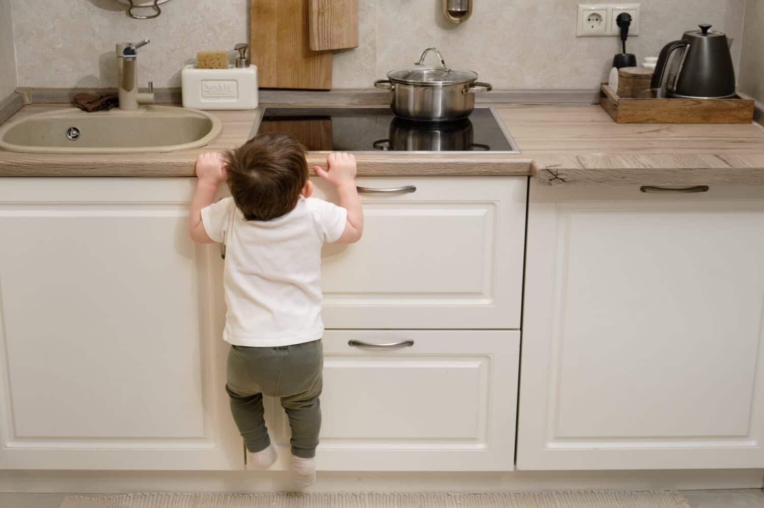 Toddler baby climbs on a hot electric stove in the home kitchen. A small child touches the surface of the stove with his hand at the risk of getting burned. Kid aged one year eight months