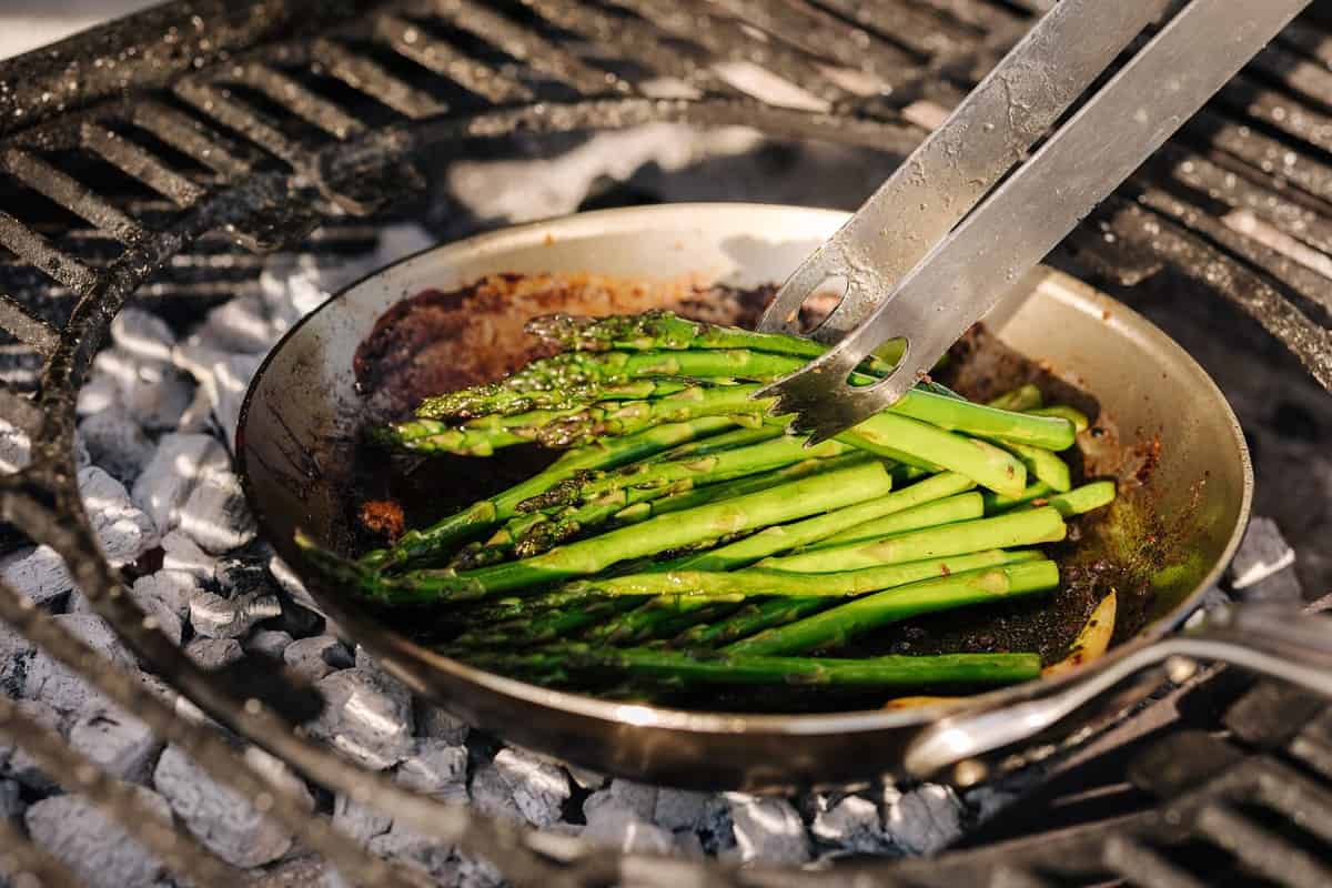Close-up of green frying asparagus outdoor on BBQ grill. Cocking vegetables on carbon steel pan. Using kitchen tongs. Hot coals