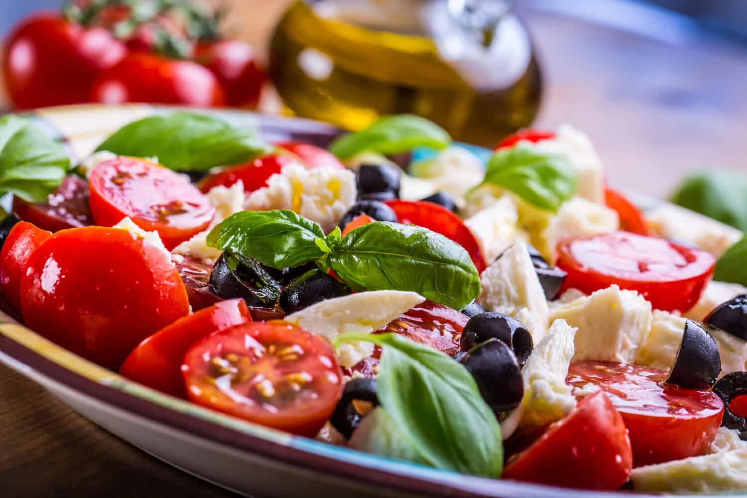 Caprese Italian or Mediterranean salad. Tomato mozzarella basil leaves black olives and olive oil on wooden table.