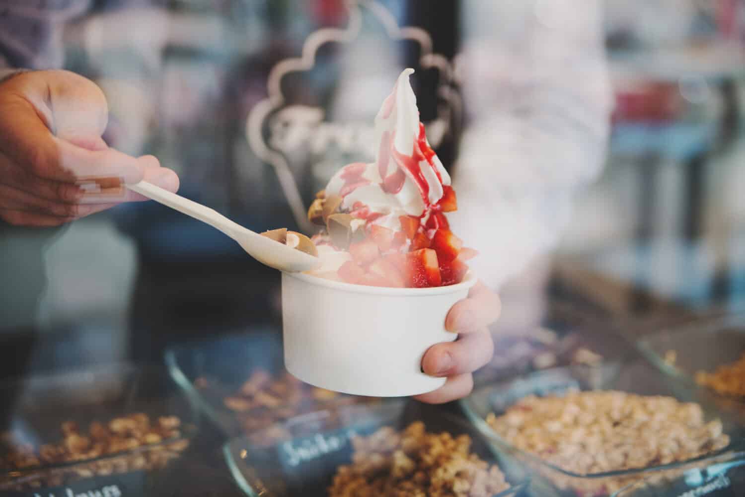 seller pours sauce on a soft frozen yoghurt in white take away cup in cafe