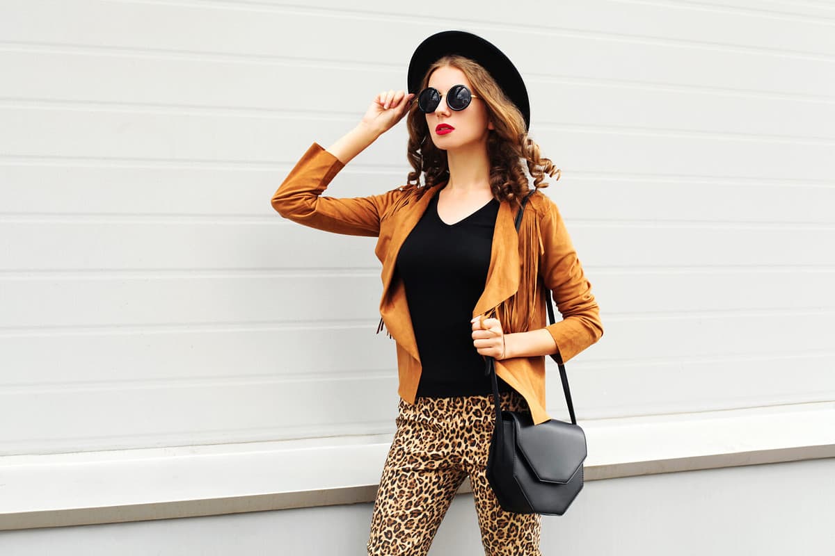 Fashion pretty woman wearing a retro elegant hat, sunglasses, brown jacket and black handbag walking in city over grey background