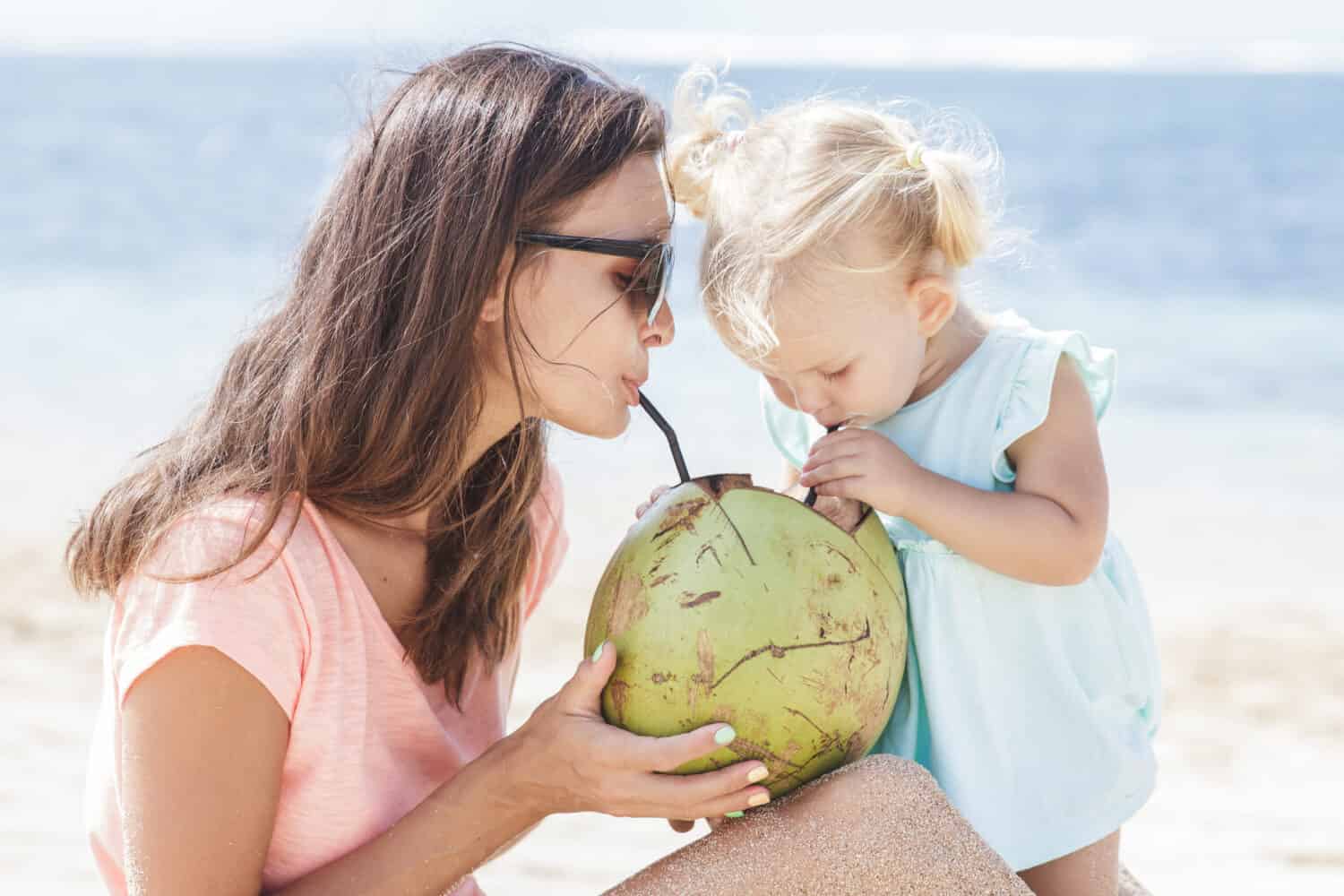 portrait of young mother sharing coconut water with her daughter while enjoying beach vacation