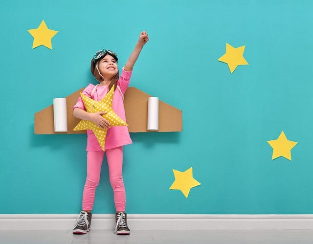 Little child girl in an astronaut costume is playing and dreaming of becoming a spaceman. Portrait of funny kid on a background of bright blue wall with yellow stars.