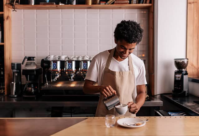 Young african man pouring milk into coffee making espresso. Professional barista preparing coffee on counter.