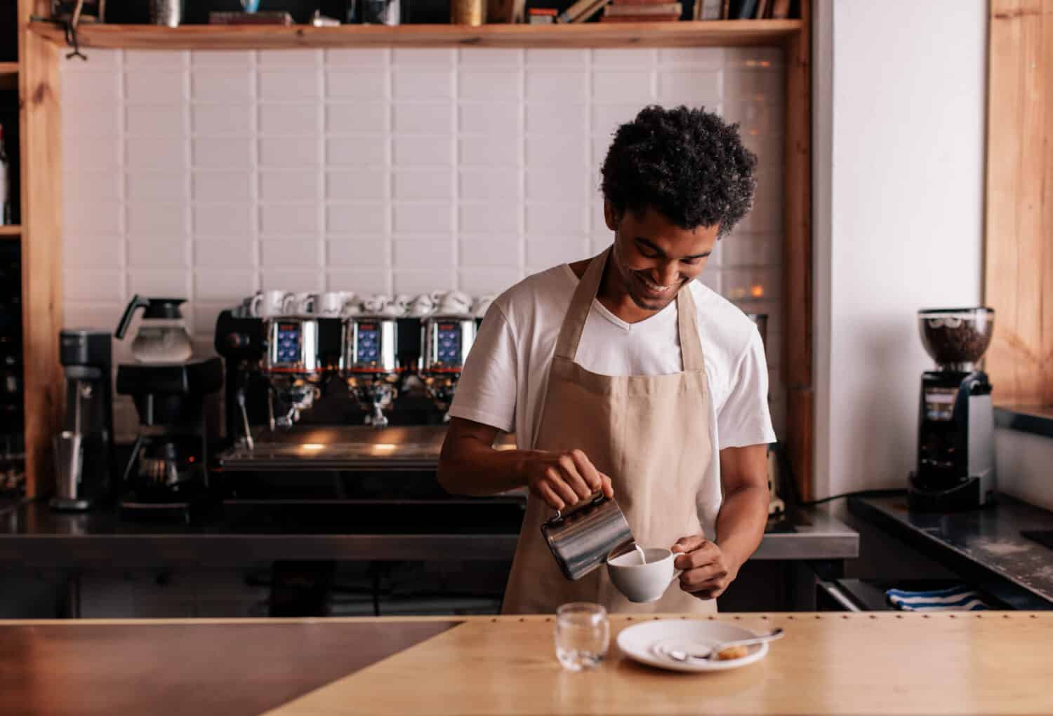 Young african man pouring milk into coffee making espresso. Professional barista preparing coffee on counter.