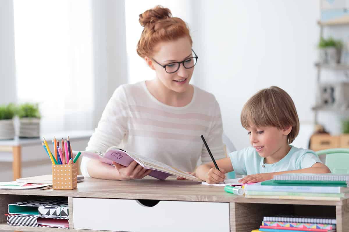 Female private tutor helping young student with homework at desk in bright child's room