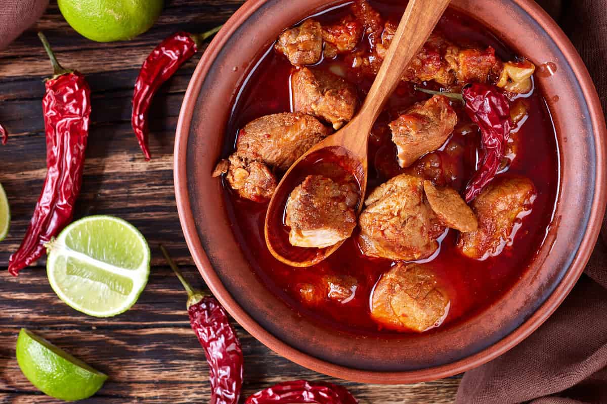 Carne Adobada or hearty Red Chile and Pork Stew in a clay bowl with spoon on an old wooden table with lime slices and dried chile, mexican cuisine, horizontal view from above, close-up, flat lay