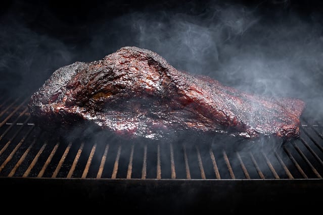 Smoke rising around a slow cooked beef brisket on the grill grates of a smoker barbecue, in a grilling concept with space for text on top and bottom