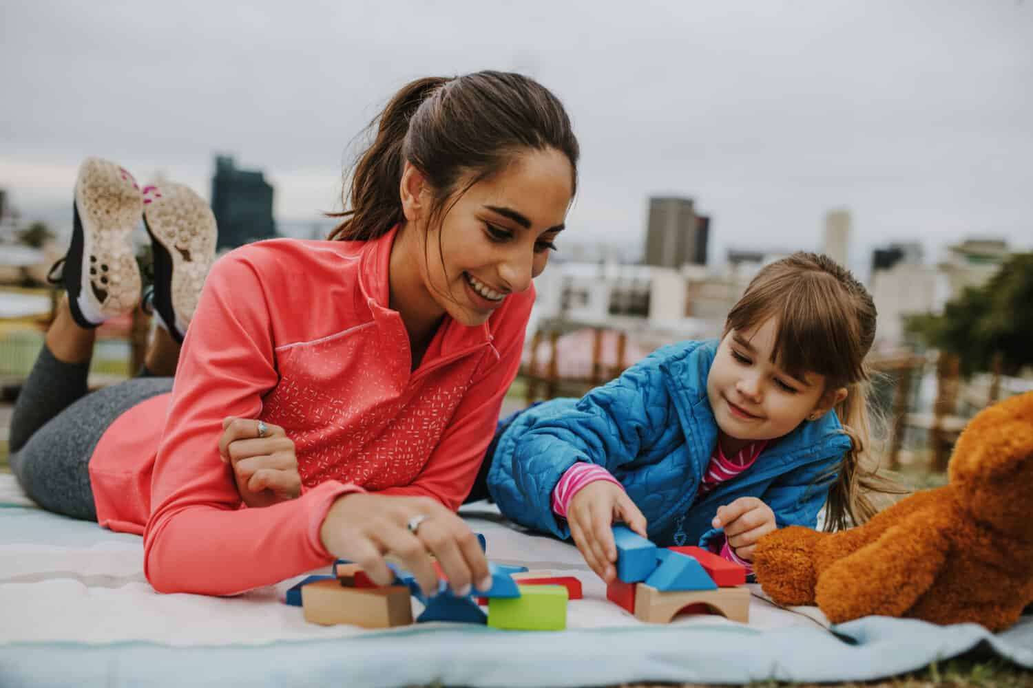 Woman playing with a small girl at the park. Girl child playing building blocks with her nanny at the park. Both lying on a plaid.