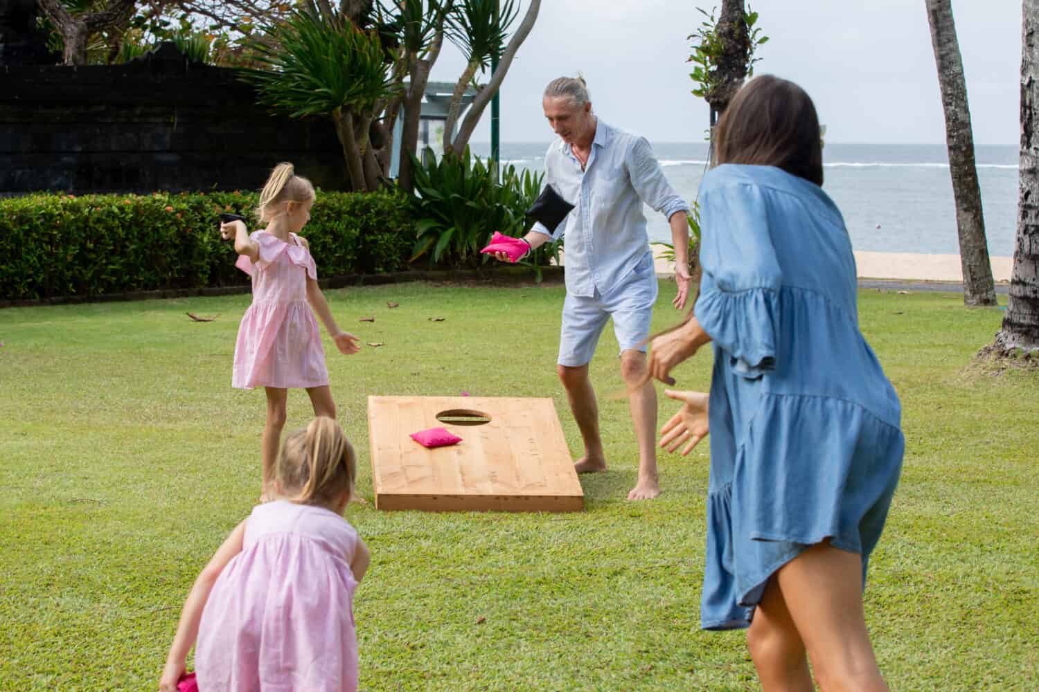 Family playing cornhole game by the sea on sunny summer day. Parents and children playing bean bag toss