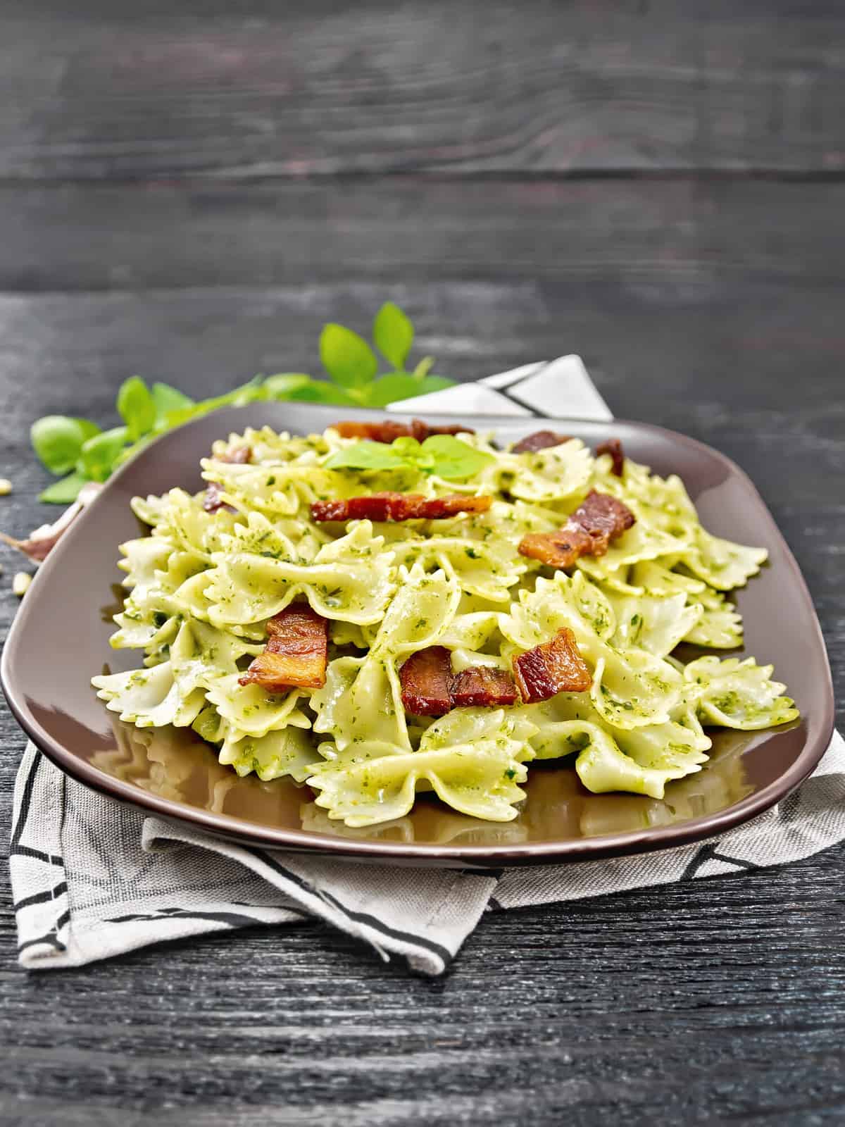 Farfalle pasta with pesto sauce, fried bacon and basil in a plate on a napkin against dark wooden board