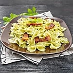 Farfalle pasta with pesto sauce, fried bacon and basil in a plate on a napkin against dark wooden board