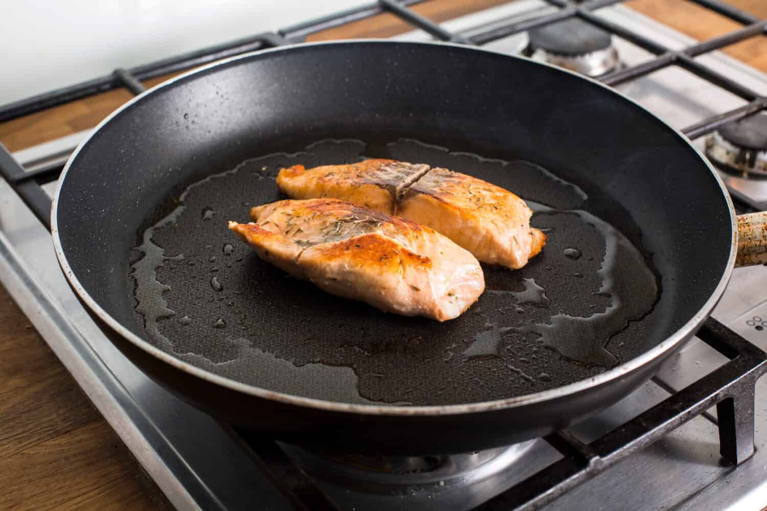 pieces of salmon cooking on a hot pan with olive oil on a gas stove.