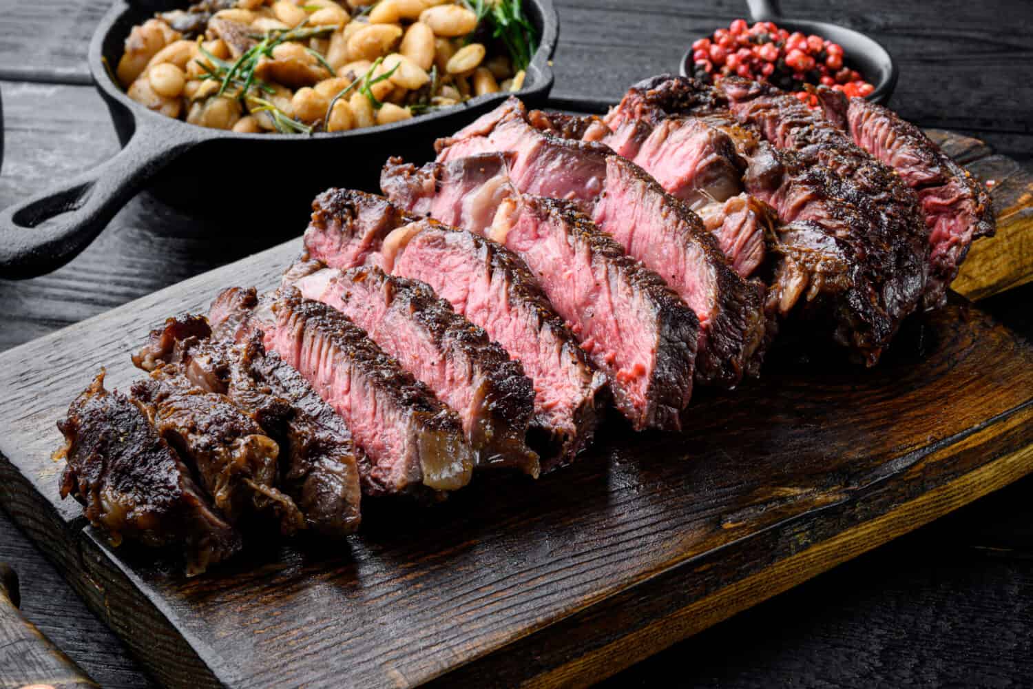 Sliced grilled meat steak Rib eye medium rare set, on wooden serving board, with white beans and rosemary in cast iron pan, on black wooden table background