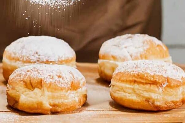 Woman prepares fresh donuts with jam in home kitchen. Cooking traditional Jewish Hanukkah sufganiyot. Hands sprinkle Berliners with powdered sugar.