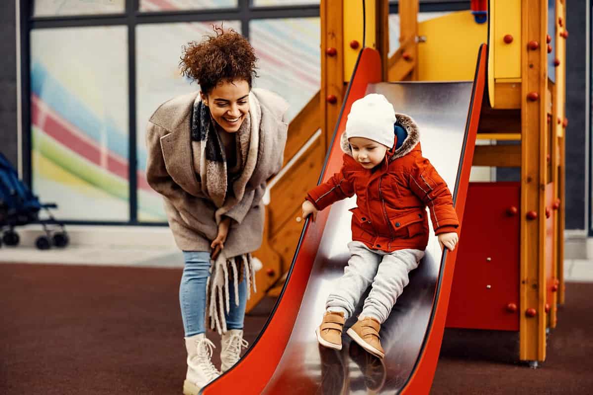 A nanny babysitting a little boy at the playground. A woman encourages a little boy on the slides.