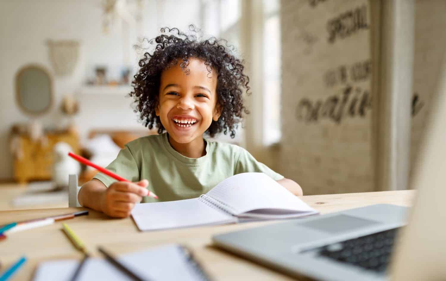 Distance education. Smiling african american child schoolboy studying online on laptop at home, sitting at table and communicating with teacher through video call on computer