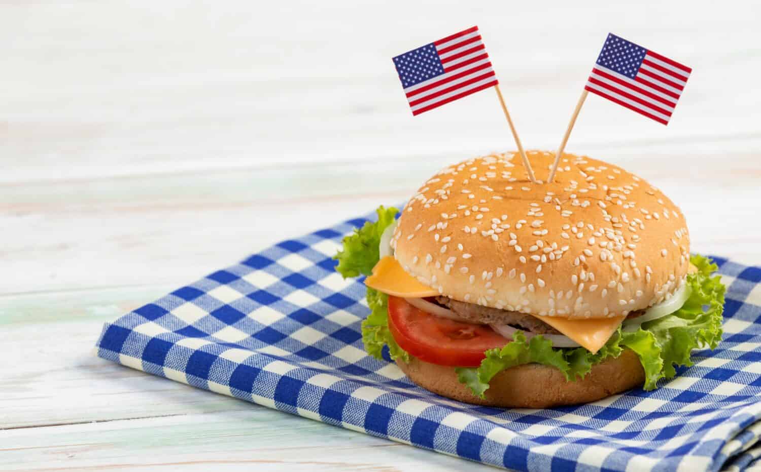 Closeup studio shot delicious tasty minced beef meat cheeseburgers fast food with two small United States of America national flag pinned on bread bun placed on plaid napkin on old retro wooden table.