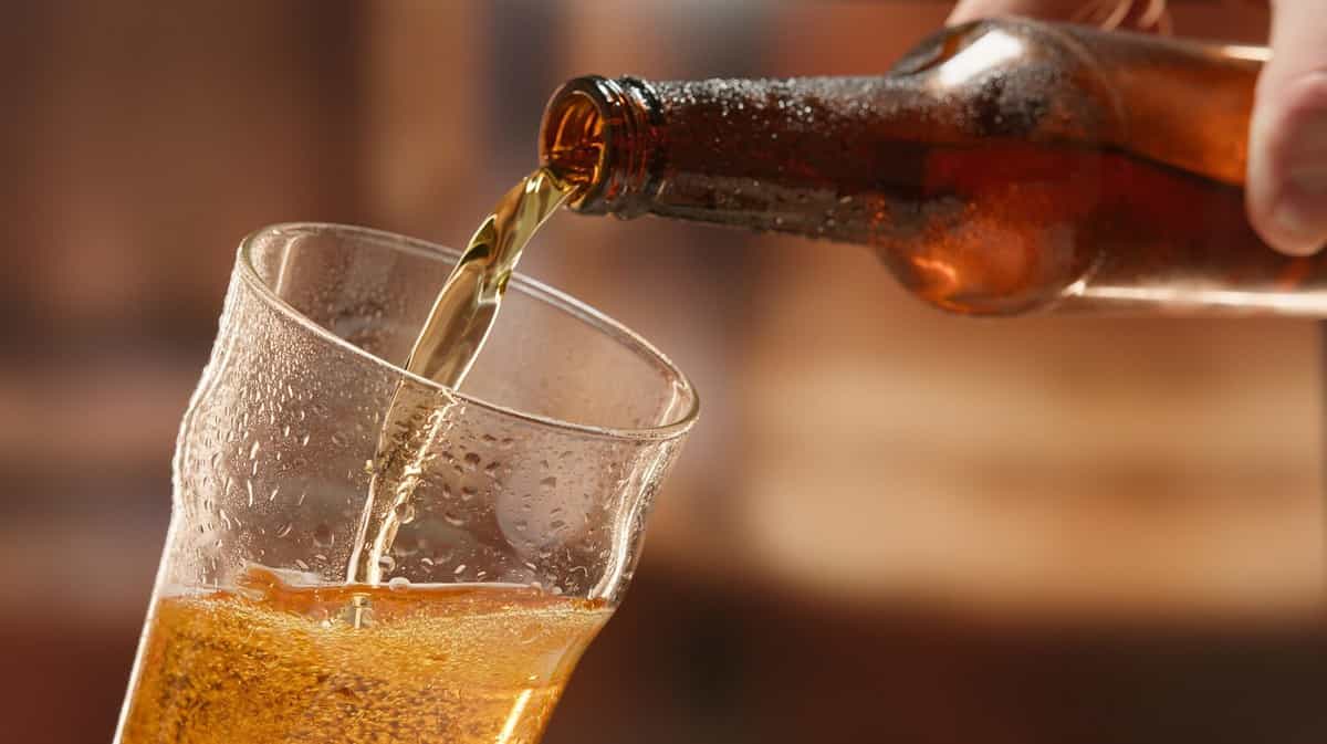 Beer is poured from dark brown bottle into beer glass. Close-up light fresh beer poured into glass steamed up from cold. Lager beer foams and pours from bottle into glass.