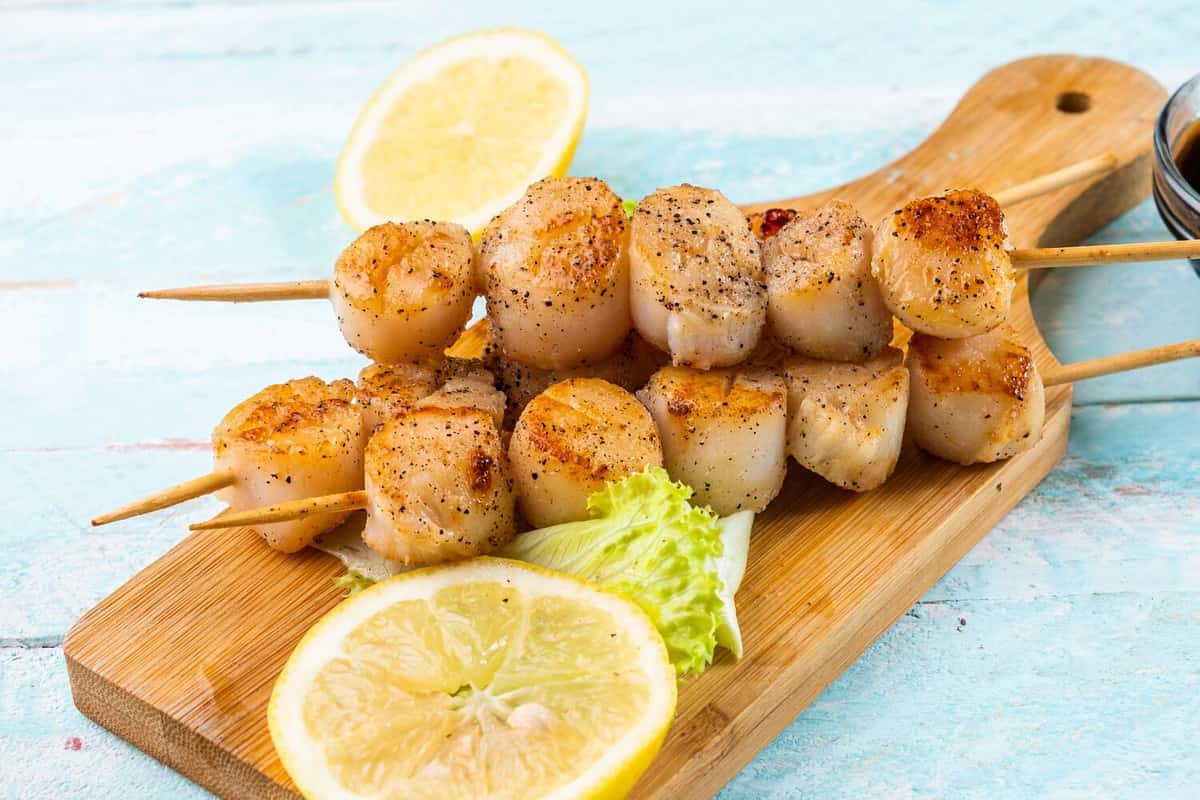 Grilled sea scallop on three skewers with lemon and spices on a wooden board. Wooden blue background.