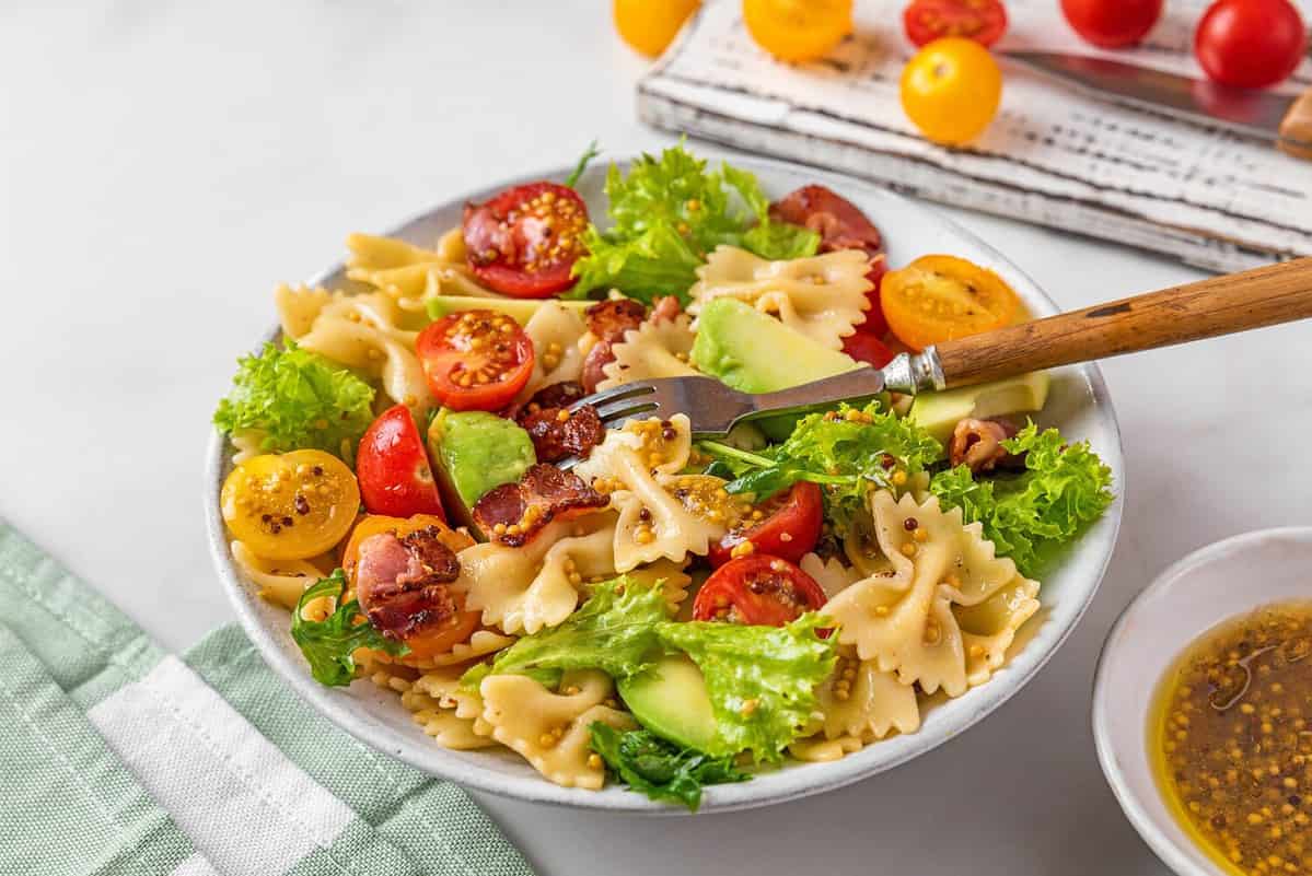 Cold summer pasta salad with bacon, tomatoes, avocado and mustard in a plate with fork on white background. Healthy diet food