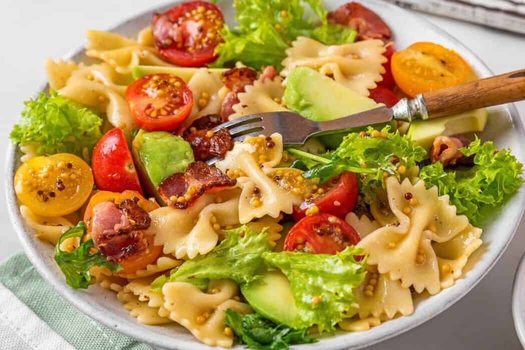 Cold summer pasta salad with bacon, tomatoes, avocado and mustard in a plate with fork on white background. Healthy diet food