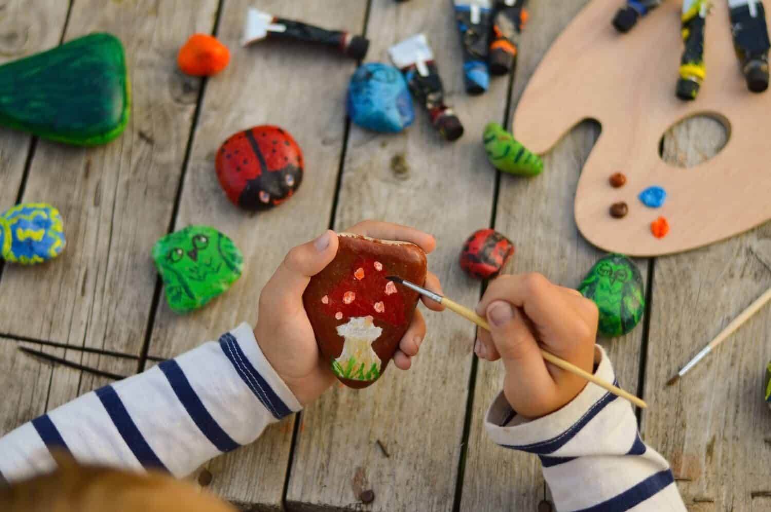 A detailed picture of a child's hands drawing a fungus on a stone with acrylic paints. Home hobbies are authentic. Artwork on stones.