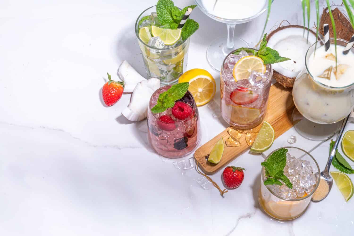 Summer cocktails drinks. Classic alcoholic long drink mocktail versions, various beverages, lemonades with berries, lime, orange, coconut, ice on white marble background