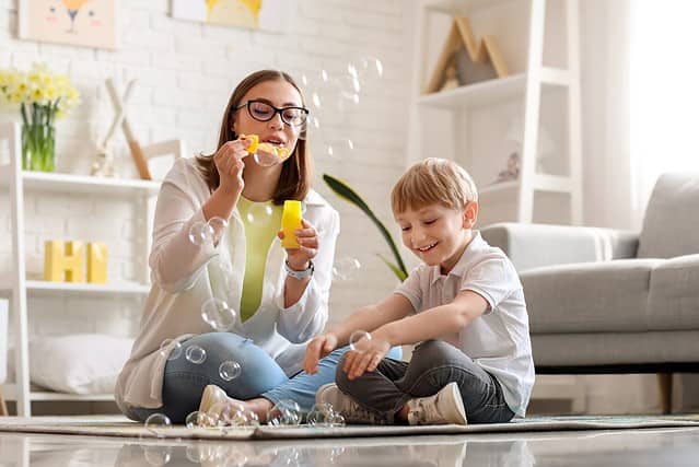 Nanny with little boy blowing soap bubbles at home