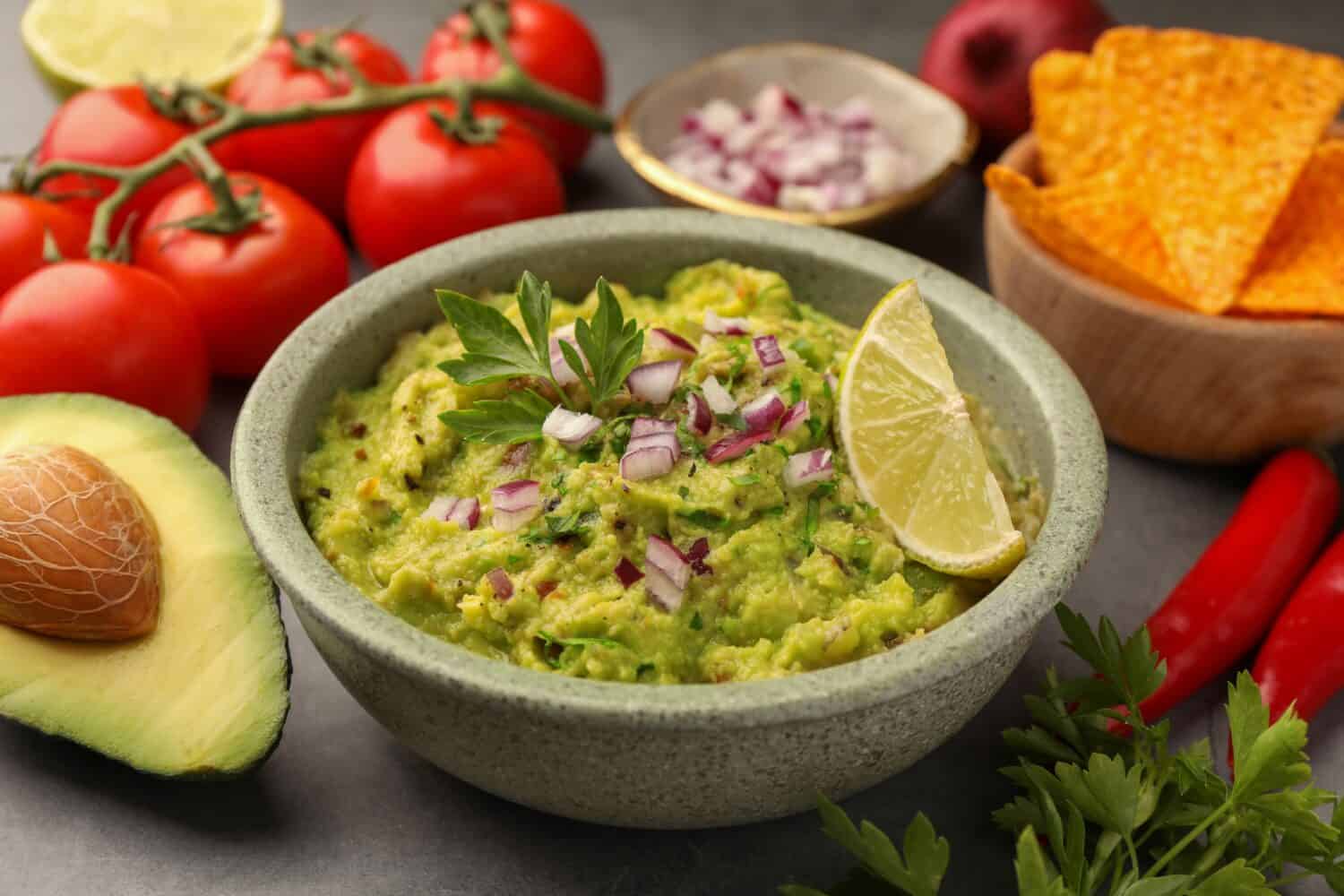 Bowl of delicious guacamole, nachos chips and ingredients on grey table, closeup