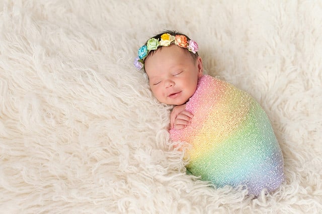 A smiling nine day old newborn baby girl bundled up in a rainbow colored swaddle. She is lying on a cream colored flokati (sheepskin) rug and wearing a crown made of roses.