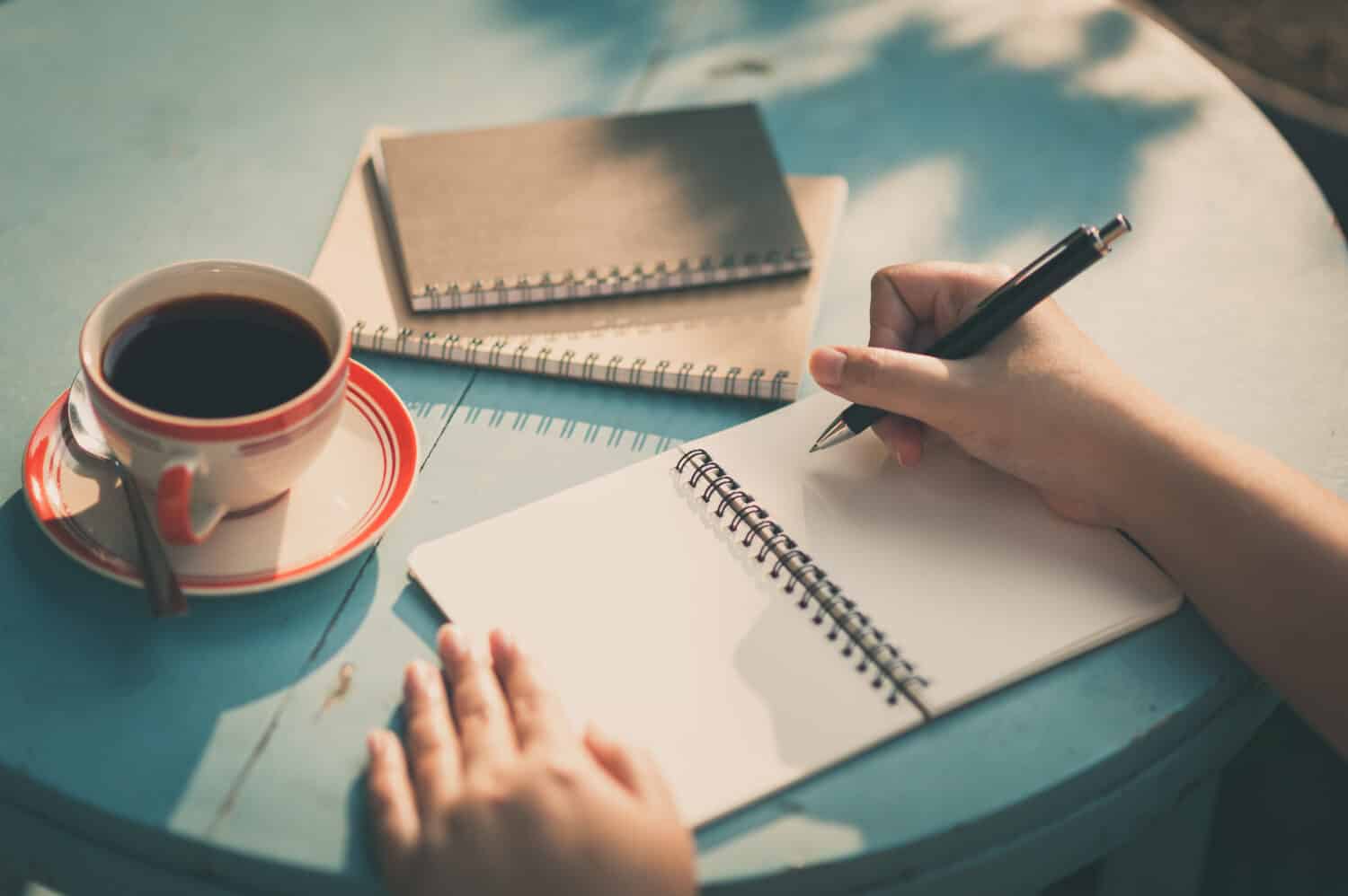 Woman right hand writing journal on small notebook at outdoor area in cafe with morning scene and vintage filter effect