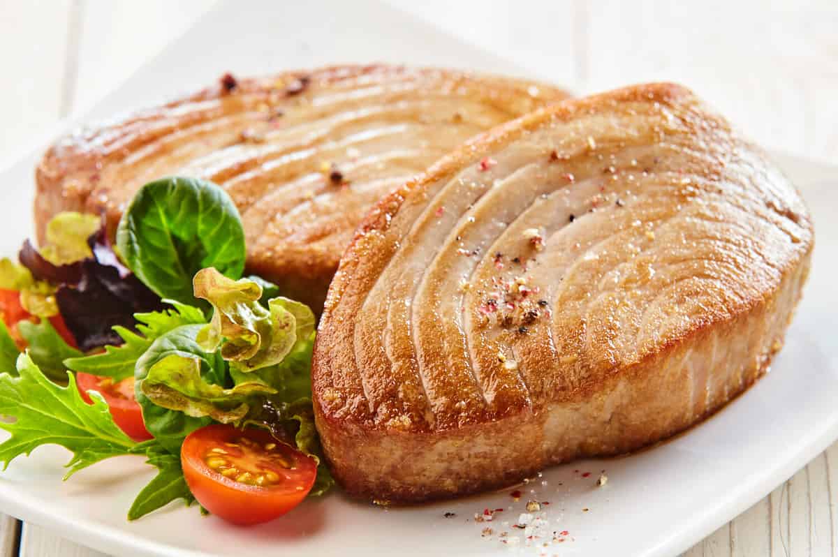 Close Up Still Life of Two Fried Tuna Steaks on Square White Plate with Seasoning and Fresh Green Salad with Tomato on Painted Wooden Table