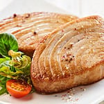 Close Up Still Life of Two Fried Tuna Steaks on Square White Plate with Seasoning and Fresh Green Salad with Tomato on Painted Wooden Table