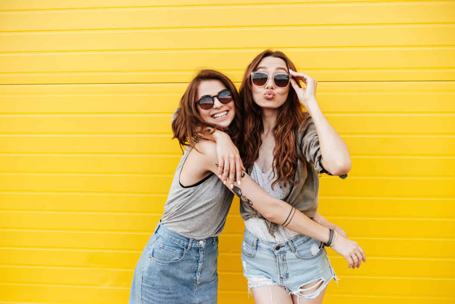 Image of two young happy women friends standing over yellow wall. Looking at camera blowing kisses.