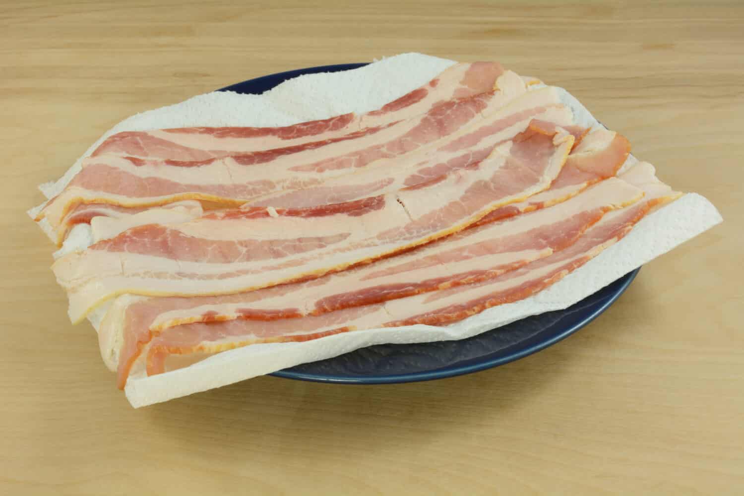 Raw bacon strips on paper towel on blue plate in preparation for microwaving