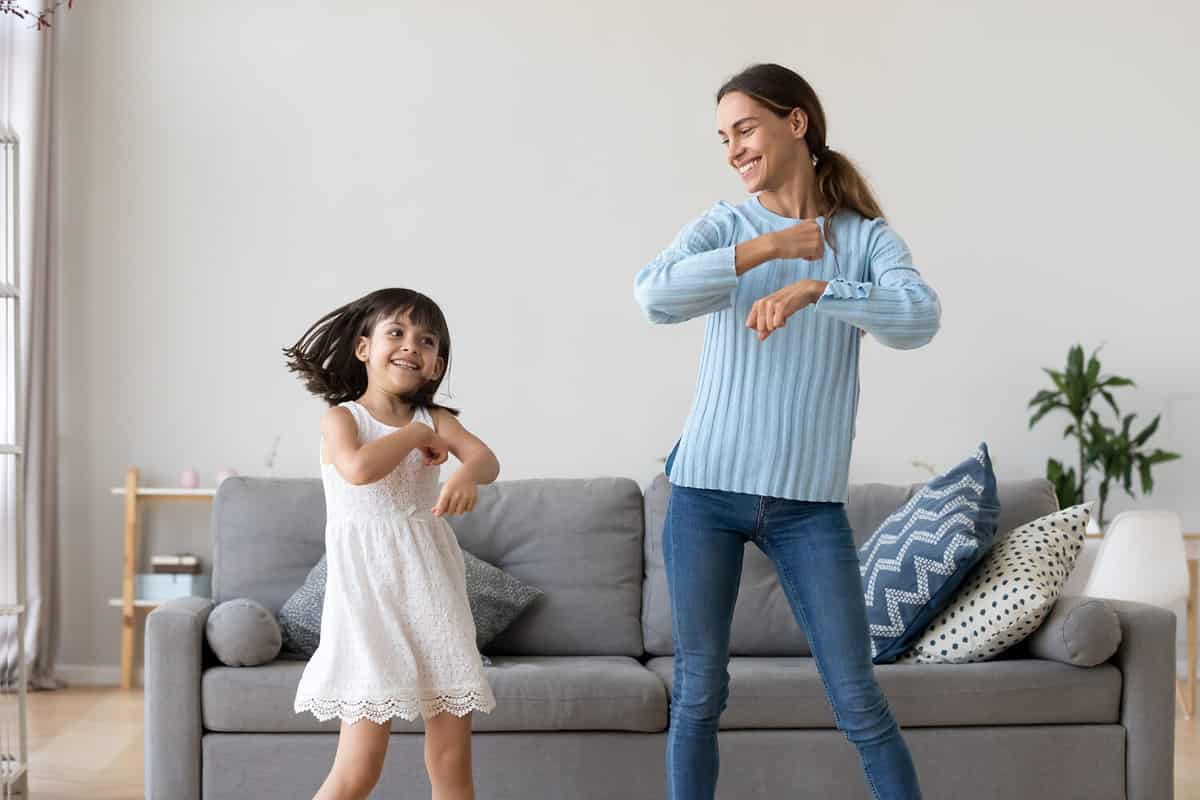Cheerful mother little daughter standing in living room at home moving dancing to favourite song together. Child have fun with elder sister nanny or loving mother active leisure and lifestyle concept