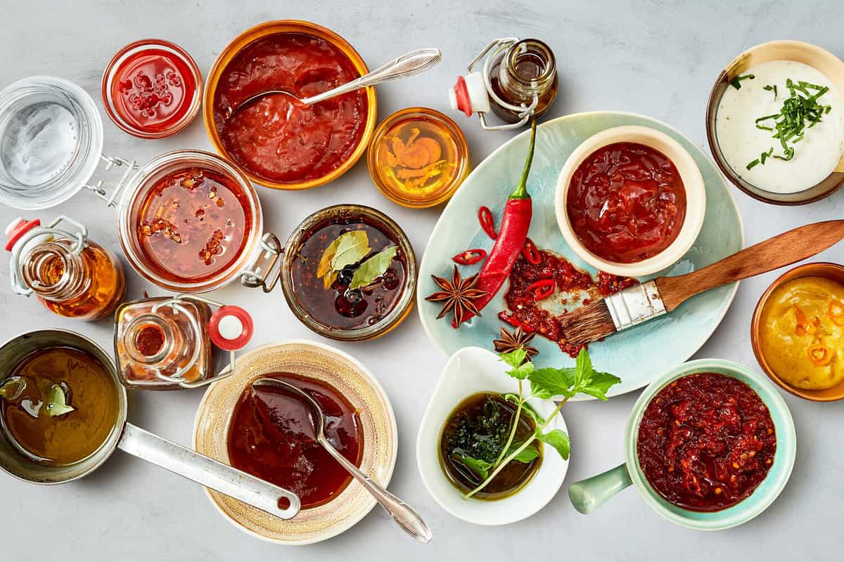 Top down view on multiple white bowls holding an assortment of bright red marinades next to plates of herbs and other ingredients