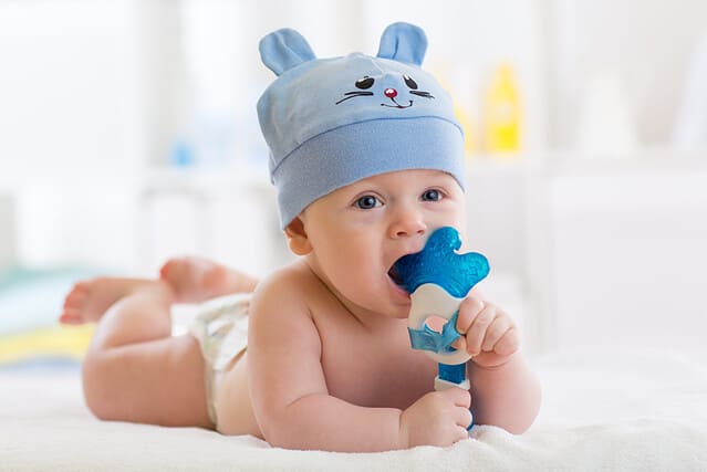 Portrait of infant baby boy weared funny hat. Kid lying on bed and playing with teething toy