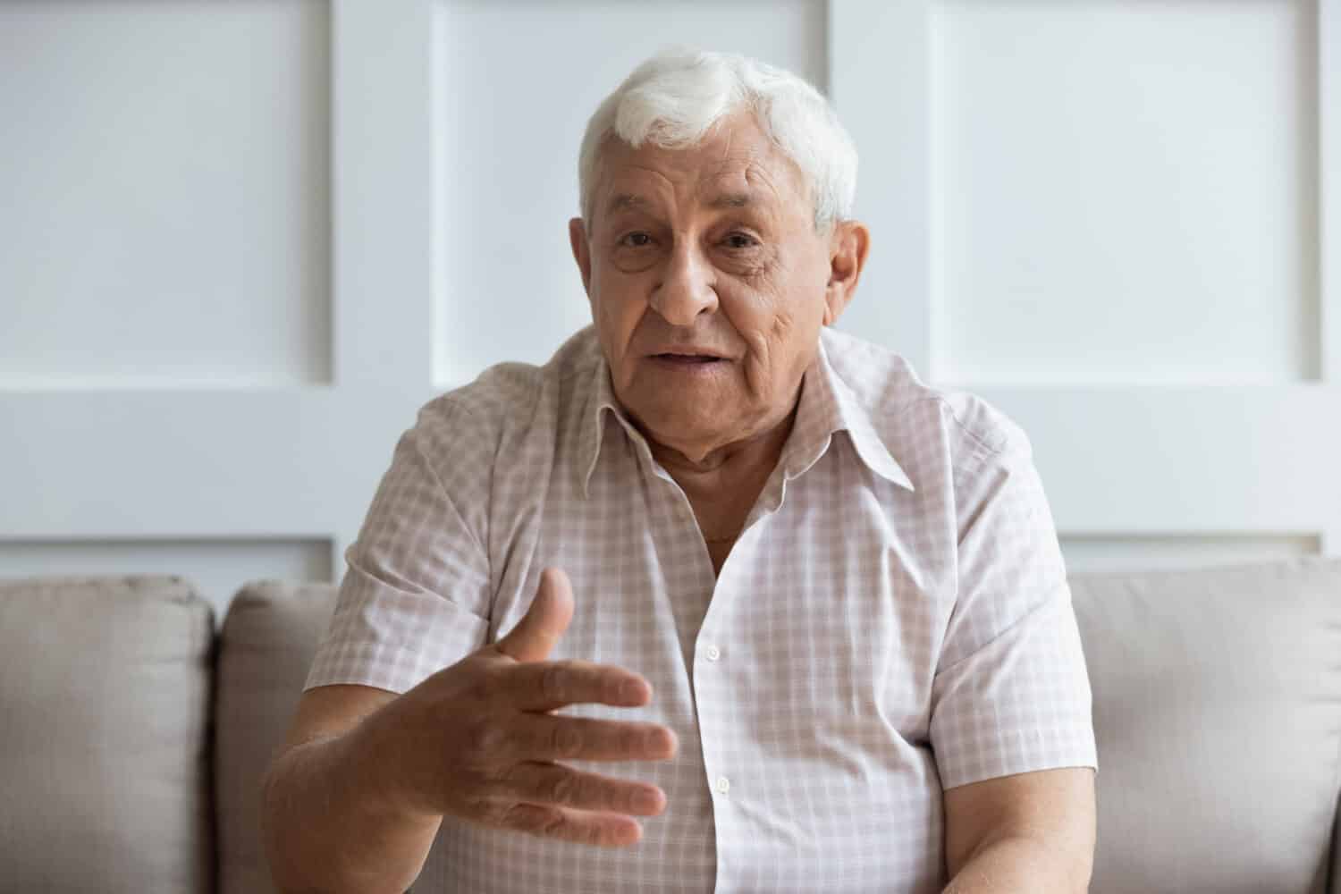 Head shot portrait serious older man looking at camera and talking, sitting on couch, grandfather making video call to grandchild on computer, mature elderly male blogger teacher speaking to webcam